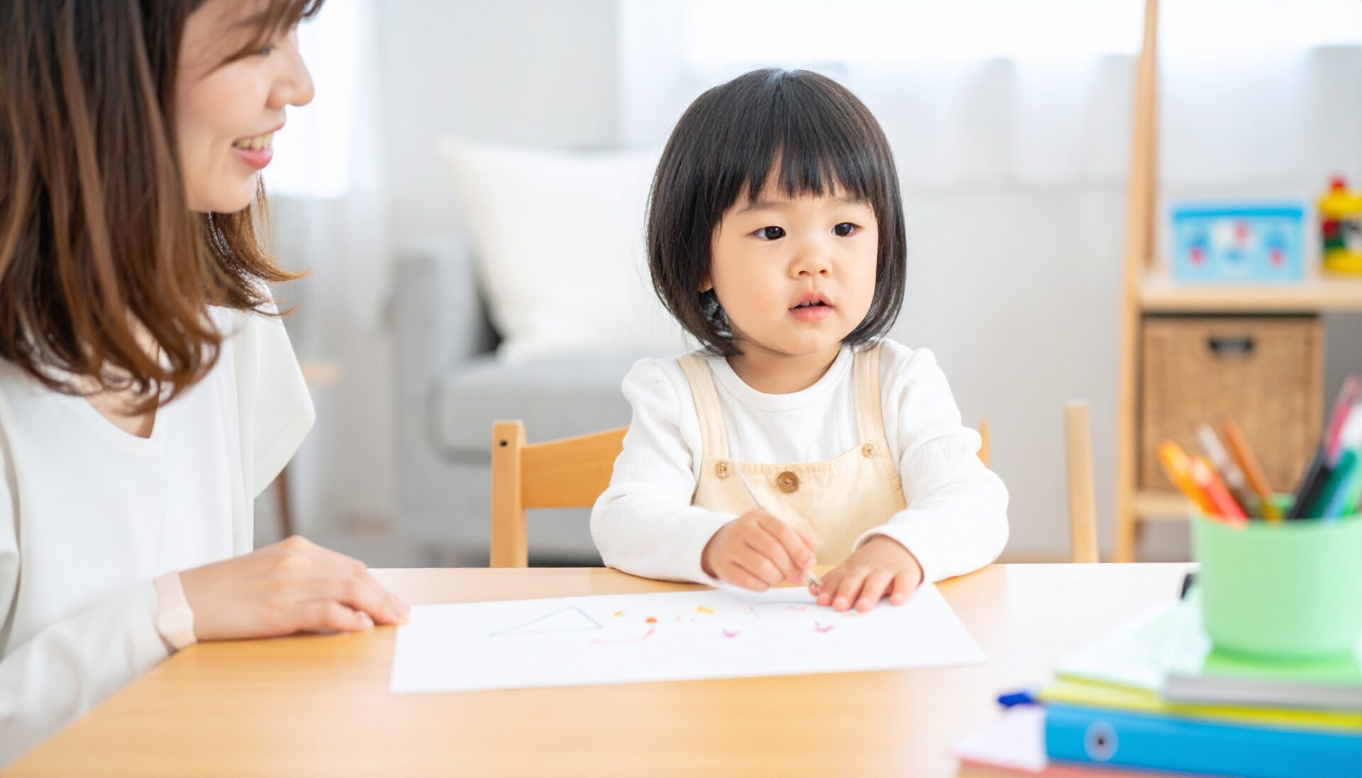 日本人の幼児が自宅のダイニングテーブルで無料の知育プリントに集中して取り組んでいる様子。横にはやさしく見守る母親。自然光の入るリビングで、カラフルな教材や文房具が整然と並ぶ。親子の笑顔と、学ぶ楽しさが伝わる暖かい雰囲気。