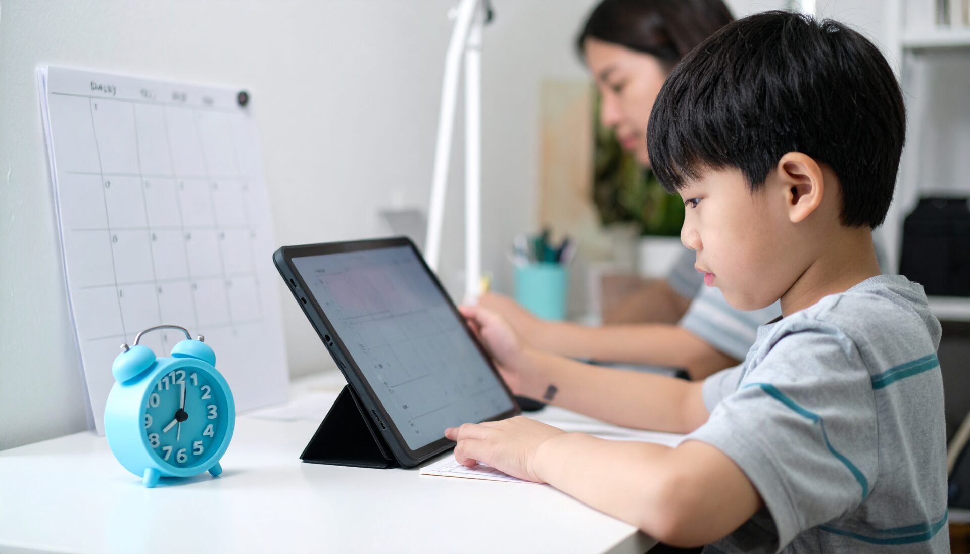 子どもの学習机に、タイマーと1日の学習スケジュール表が貼られており、子どもが自分で時間を見ながらタブレット学習している。親は別の作業中。