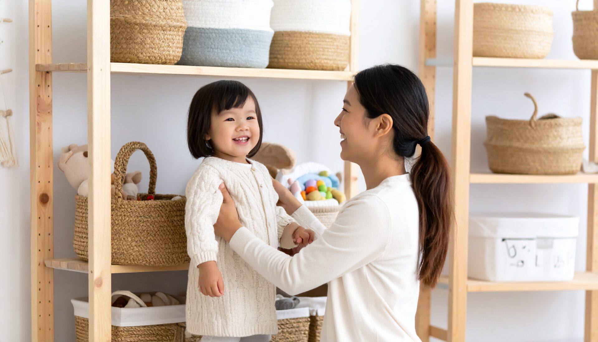 A Japanese toddler has just finished tidying up toys and is proudly smiling or saying “I did it!” while a parent looks on warmly, sharing the joy. The room features natural materials like wooden shelves and fabric bins, creating a calm, organized environment. The scene highlights the emotional reward and developmental value of encouraging independence through daily routines.
